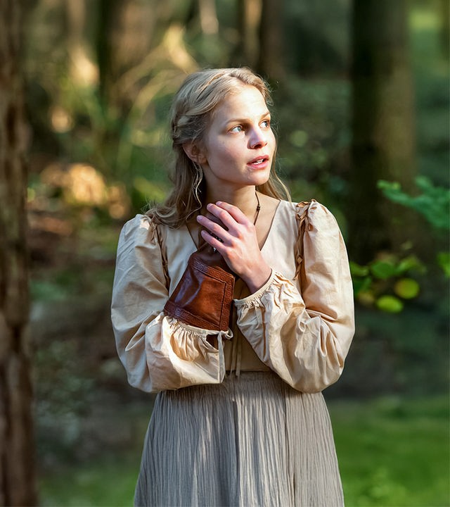 Constanze, verkörpert von Sinja Dieks, steht in Bauernkleidung in einem Wald. Links von ihr ist ein dicker Baumstamm. Contanze hat ihre Hände vor ihrer Brust verchränkt. Die rechte Hand steckt in einem ledernen Handschuh, mit dem man sonst Wildvögel auf der Hand hält. Constanze schaut aufmerksam und leicht angespannt nach rechts.