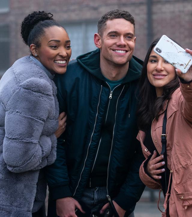 Billy (Tom Brittney) steht zwischen zwei Frauen, von denen eine ein Handy hochhält, um ein Selfie zu machen. Alle lächeln in die Kamera. 