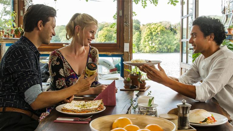 "Matze, Kebab und Sauerkraut": Noah (Franz Dinda), Charlottes (Christine Eixenberger) und Hakim (Omar El-Saeidi) sitzen gemeinsam am Tisch. Hakim hält Charlotte einen Teller hin, im Hintergrund glitzert der See.