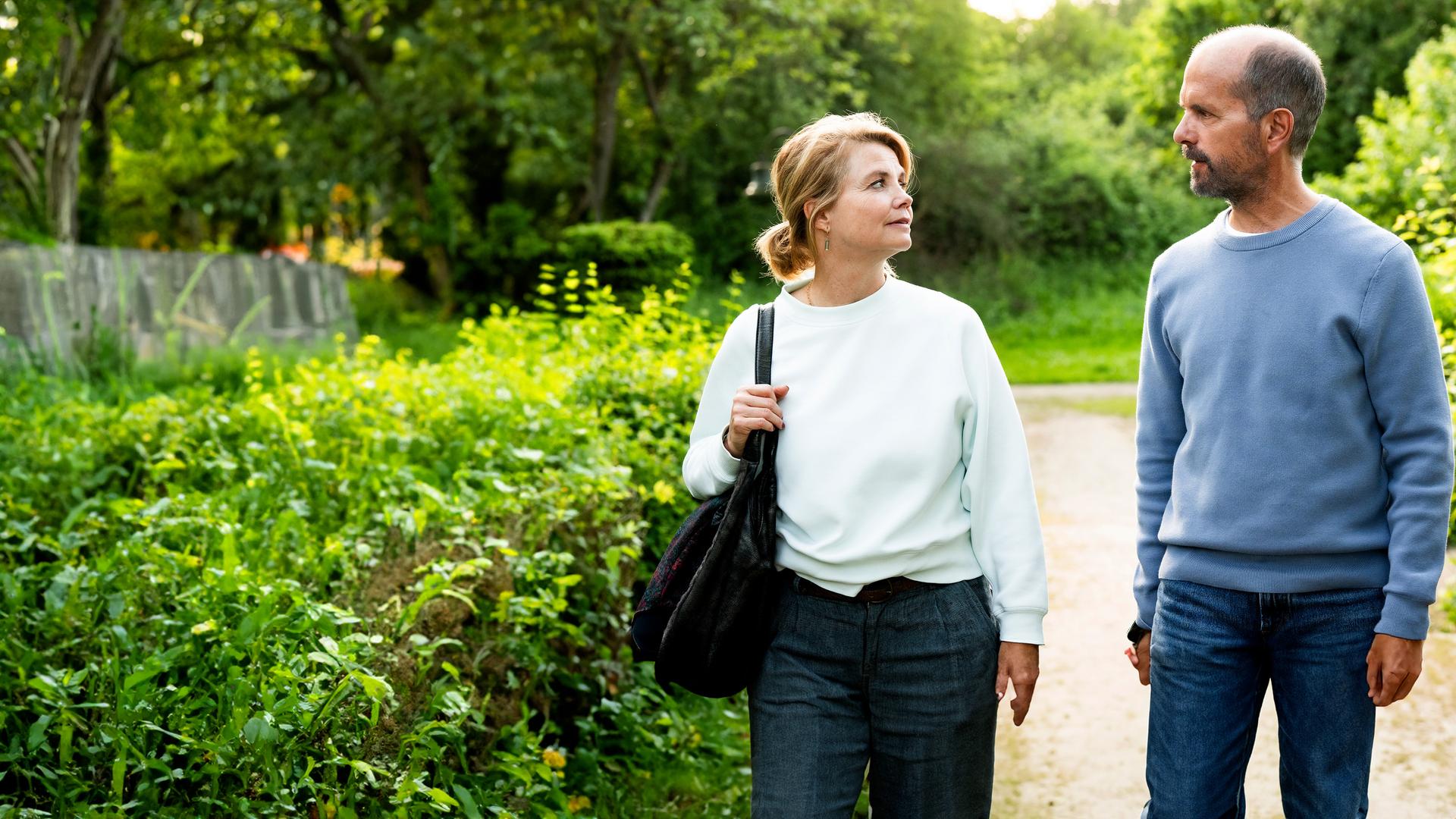 Erik (Christoph Maria Herbst) und Anne (Annette Frier) spazieren an einem sonnigen Tag über einen Feldweg und sehen sich dabei an.