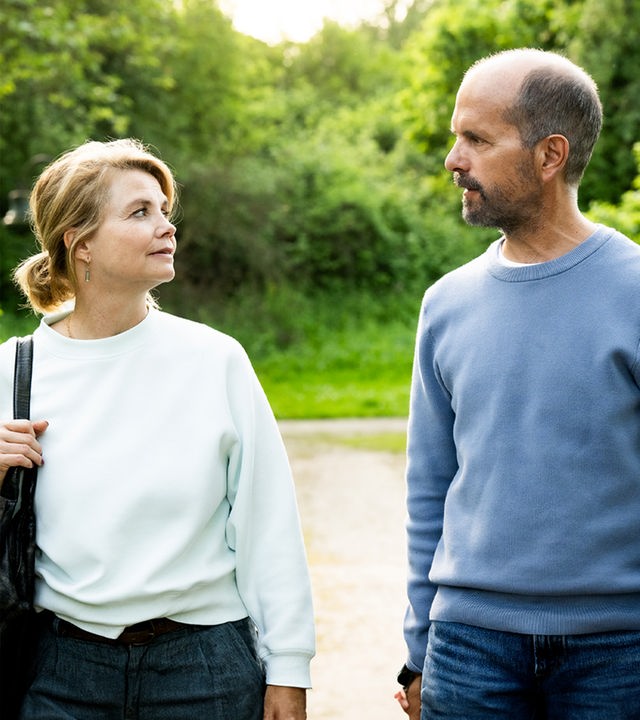 Erik (Christoph Maria Herbst) und Anne (Annette Frier) spazieren an einem sonnigen Tag über einen Feldweg und sehen sich dabei an.