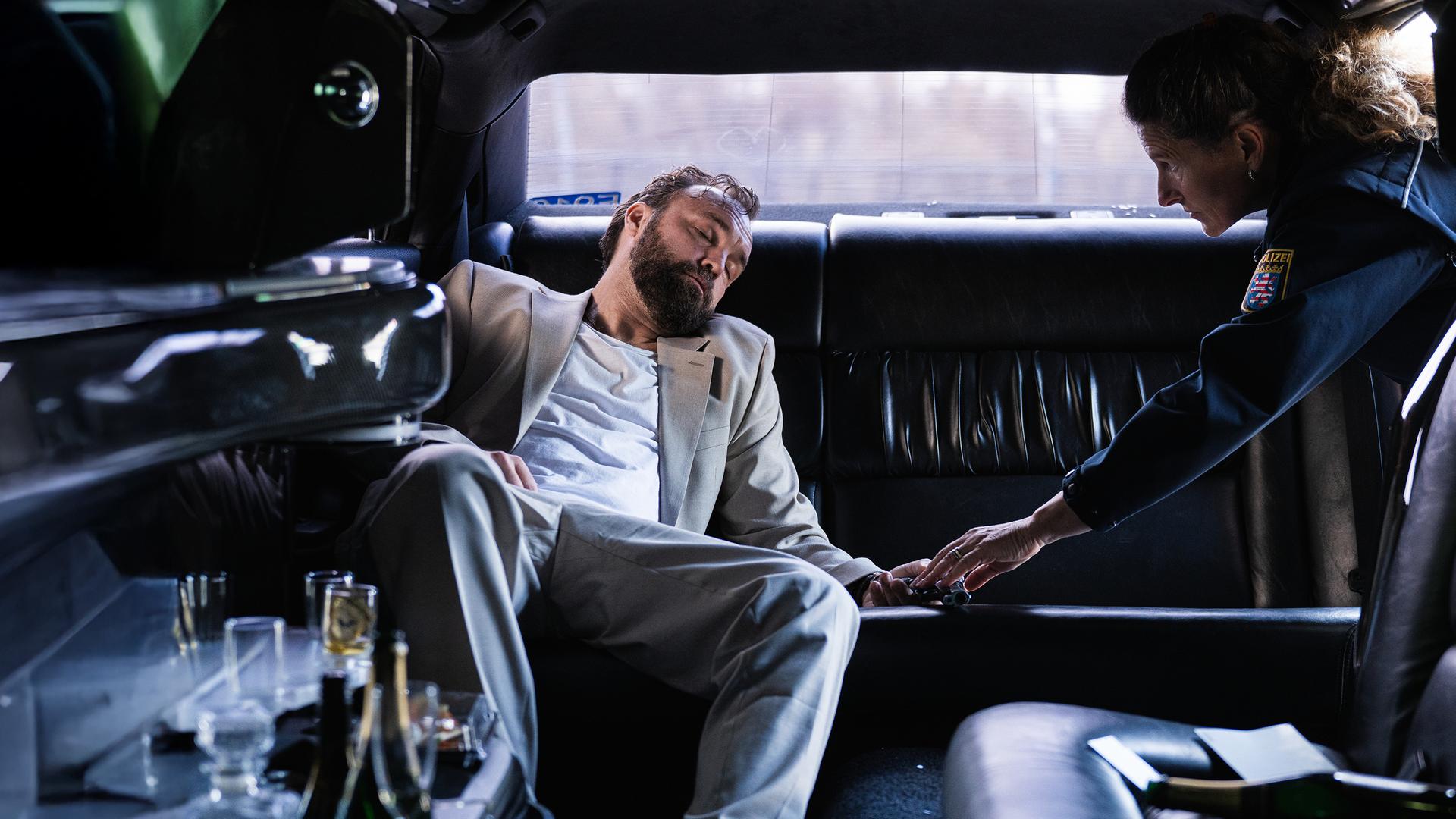 In einer dunklen Limousine liegt Mark Liffert (Isaak Dentler) zusammengesunken auf der Rückbank. Er trägt einen hellen Anzug, sein Kopf ist zur Seite geneigt, die Augen geschlossen. Neben ihm beugt sich eine Polizistin (Komparsin) vor und überprüft vorsichtig seine Hand. Leere Gläser und Flaschen stehen in der Bar der Limousine, eine weitere Flasche liegt auf dem Boden. Die Szene wirkt kühl beleuchtet und vermittelt den Eindruck einer außer Kontrolle geratenen Partynacht.