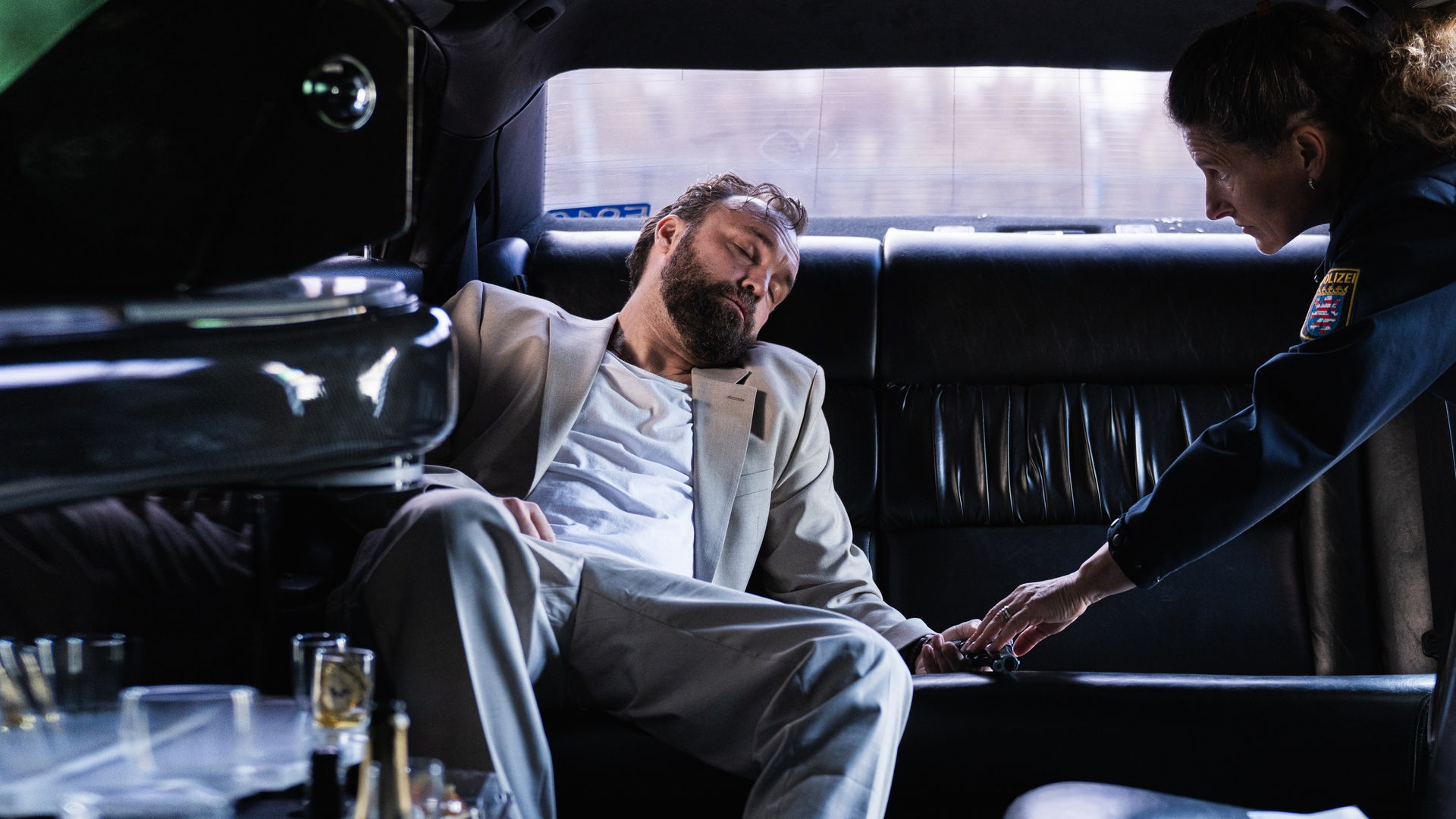 In einer dunklen Limousine liegt Mark Liffert (Isaak Dentler) zusammengesunken auf der Rückbank. Er trägt einen hellen Anzug, sein Kopf ist zur Seite geneigt, die Augen geschlossen. Neben ihm beugt sich eine Polizistin (Komparsin) vor und überprüft vorsichtig seine Hand. Leere Gläser und Flaschen stehen in der Bar der Limousine, eine weitere Flasche liegt auf dem Boden. Die Szene wirkt kühl beleuchtet und vermittelt den Eindruck einer außer Kontrolle geratenen Partynacht.