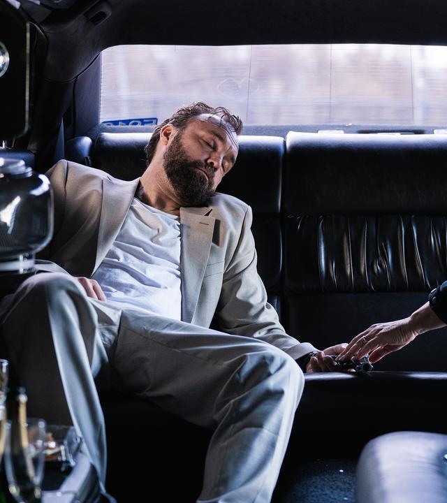 In einer dunklen Limousine liegt Mark Liffert (Isaak Dentler) zusammengesunken auf der Rückbank. Er trägt einen hellen Anzug, sein Kopf ist zur Seite geneigt, die Augen geschlossen. Neben ihm beugt sich eine Polizistin (Komparsin) vor und überprüft vorsichtig seine Hand. Leere Gläser und Flaschen stehen in der Bar der Limousine, eine weitere Flasche liegt auf dem Boden. Die Szene wirkt kühl beleuchtet und vermittelt den Eindruck einer außer Kontrolle geratenen Partynacht.