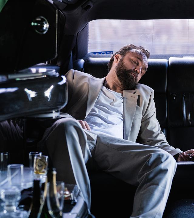 In einer dunklen Limousine liegt Mark Liffert (Isaak Dentler) zusammengesunken auf der Rückbank. Er trägt einen hellen Anzug, sein Kopf ist zur Seite geneigt, die Augen geschlossen. Neben ihm beugt sich eine Polizistin (Komparsin) vor und überprüft vorsichtig seine Hand. Leere Gläser und Flaschen stehen in der Bar der Limousine, eine weitere Flasche liegt auf dem Boden. Die Szene wirkt kühl beleuchtet und vermittelt den Eindruck einer außer Kontrolle geratenen Partynacht.