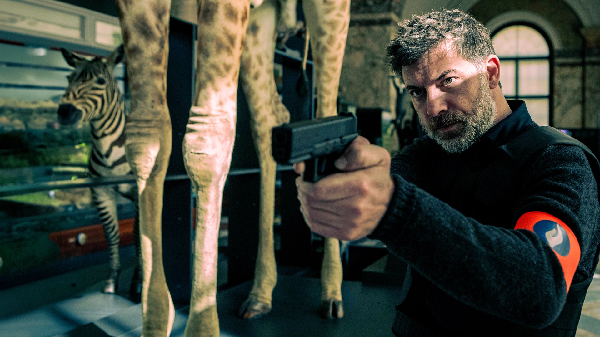 Kommissar Nick Cafmeyer (Geert van Rampelberg) steht mit gezogener Waffe in einem naturhistorischen Museum; neben ihm sind die Beine einer ausgestopften Giraffe und eines Zebras zu sehen.