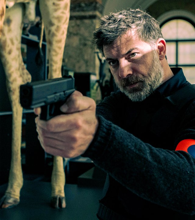 Kommissar Nick Cafmeyer (Geert van Rampelberg) steht mit gezogener Waffe in einem naturhistorischen Museum; neben ihm sind die Beine einer ausgestopften Giraffe und eines Zebras zu sehen.