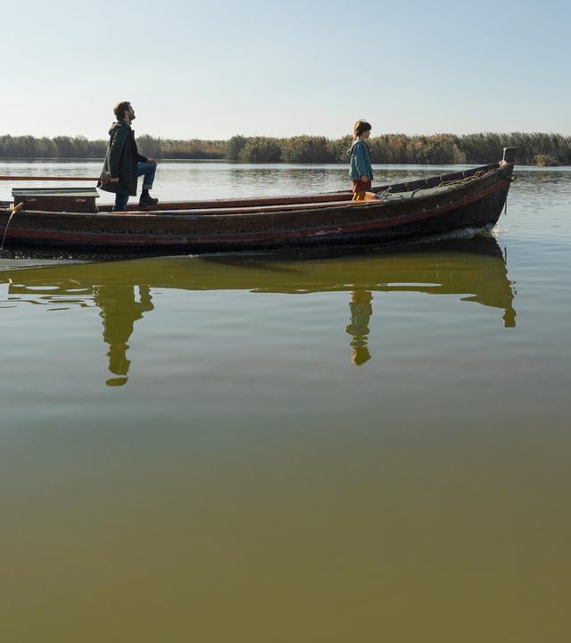 Ricardo (Raúl Arévalo) und Julia (Daniela Casas) stehen mit etwas Abstand hintereinander in langem Boot auf ruhigem Gewässer