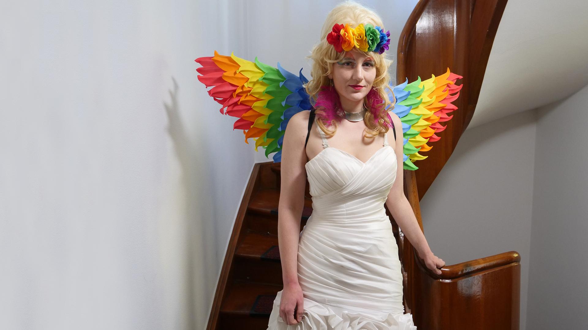 Schwester Micha steht vor einer Treppe mit einem Kirchenfenster. Sie trägt ein weißes Kleid, eine blonde Perücke und Flügel in Regenbogenfarben.