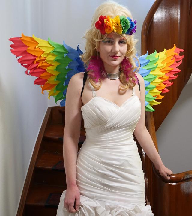 Schwester Micha steht vor einer Treppe mit einem Kirchenfenster. Sie trägt ein weißes Kleid, eine blonde Perücke und Flügel in Regenbogenfarben.
