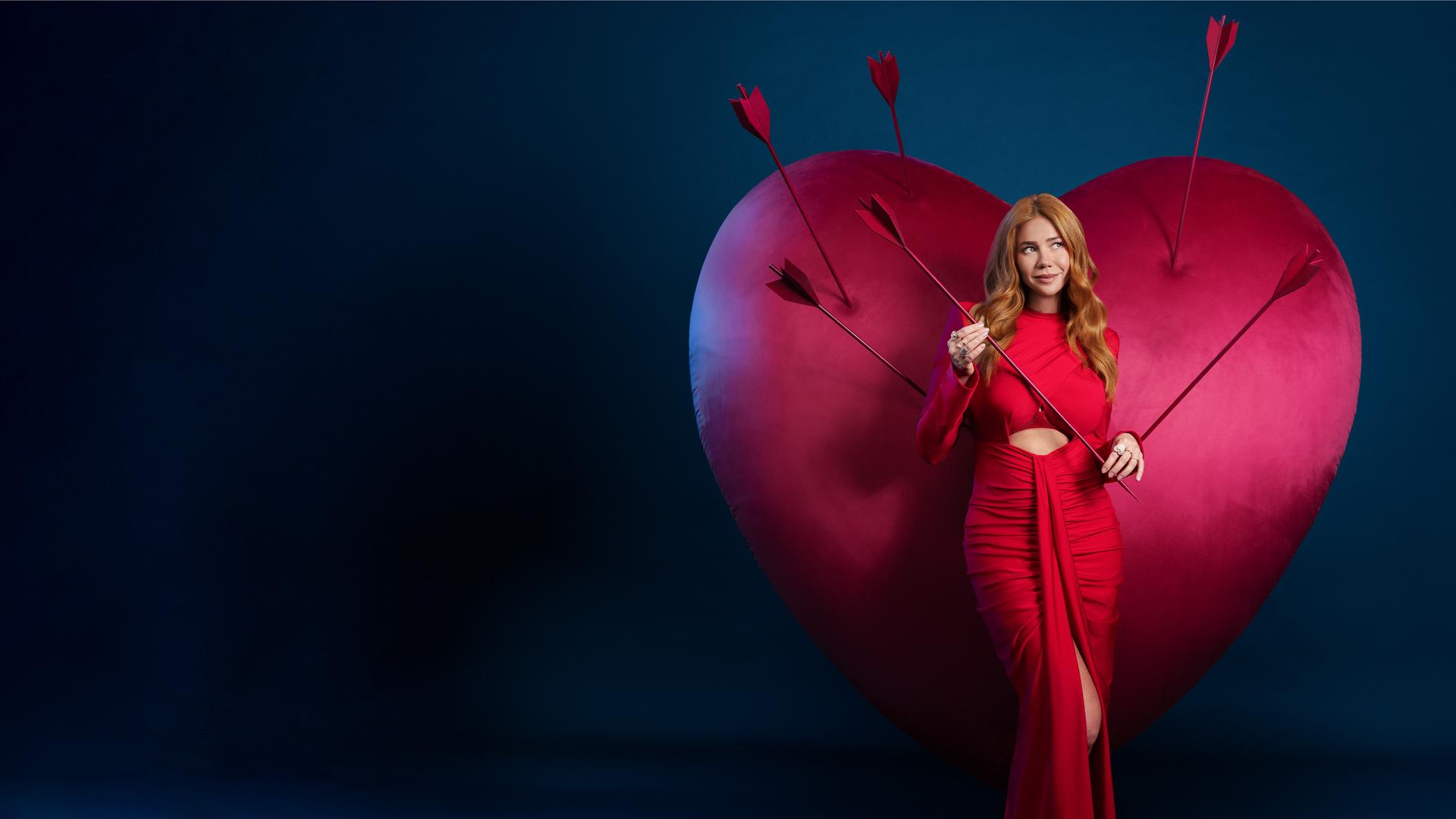 Ivy (Palina Rojinski) steht in einem langen roten Kleid vor einem großen roten Samtherz in dem einige Pfeile stecken. Sie hält einen Pfeil in ihren Händen.