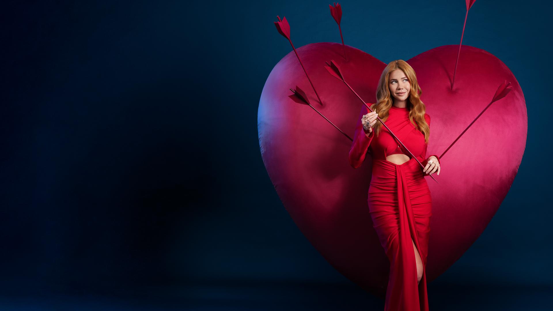 Ivy (Palina Rojinski) steht in einem langen roten Kleid vor einem großen roten Samtherz in dem einige Pfeile stecken. Sie hält einen Pfeil in ihren Händen.
