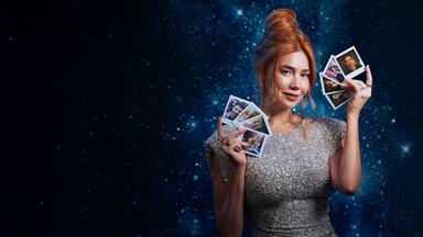 Ivy (Palina Rojinski) steht in einem langen roten Kleid vor einem großen roten Samtherz in dem einige Pfeile stecken. Sie hält einen Pfeil in ihren Händen.