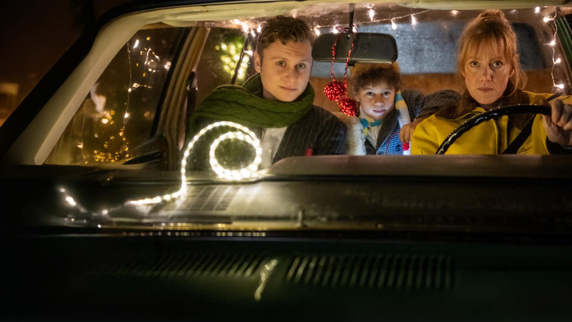 "Nelly und das Weihnachtswunder": Sonny (Rafael Gareisen), Nick (Joah Lion Goetze) und Nelly (Anna Schudt) sitzen in einem Auto, das mit Lichtergirlanden im Inneren geschmückt ist. Nelly sitzt am Steuer, Sonny sitzt neben ihr, Nick sitzt hinter den beiden und hat einen Arm auf Nellys Rückenlehne gelegt. Alle schauen interessiert nach vorne.