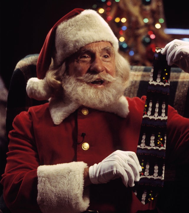 Nick (Nigel Hawthorne) sitzt in einem karierten Sessel und hält mit beiden Händen eine weihnachtliche Krawatte hoch. Er trägt eine Nikolaus-Kostüm und lacht.