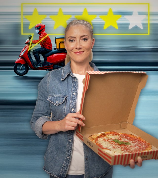 Pia Osterhaus steht in einer Großküche und hält eine fertige Pizza im Karton in der Hand. Sie schaut lächelnd in die Kamera. Im Hintergrund Im Hintergrund fährt ein Mopedfahrer mit Lieferung.  