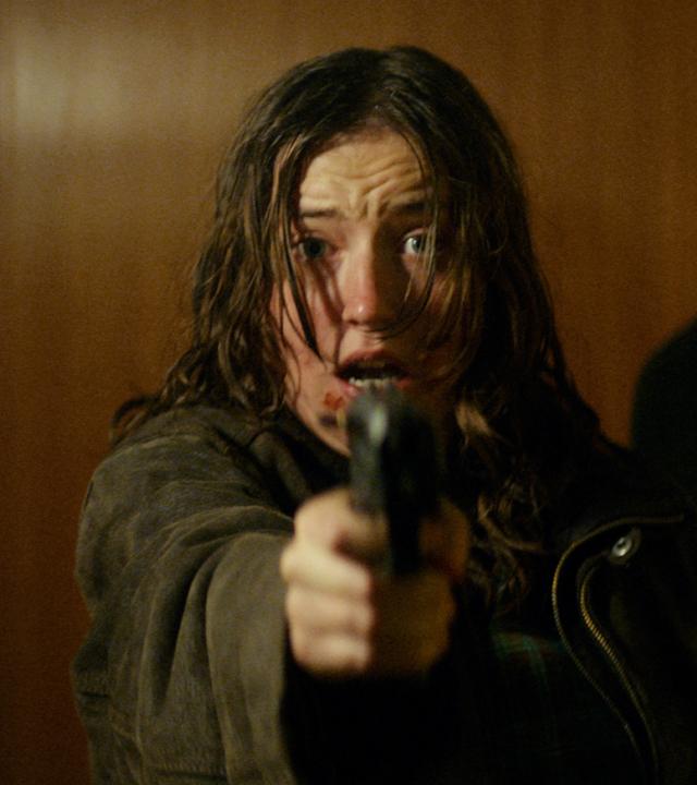 mma (Fanny Leander Bornedal) hält eine Pistole in der Hand und zielt nach vorne. Sie hat leichte Verletzungen im Gesicht und sieht ängstlich aus. Hinter ihr steht Martin (Nikolaj Coster-Waldau), auch er sieht erschrocken nach vorn.