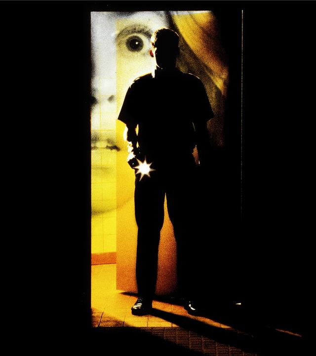 Plakatmotiv zum Film "Nightwatch - Nachtwache" mit Nikolaj Coster-Waldau. "Nightwatch - Nachtwache": Montage: In einem dunklen Raum ist eine Tür geöffnet, durch die Licht einfällt. In der Tür steht Martin (Nikolaj Coster-Waldau) mit einer Taschenlampe. Hinter der Tür ist der entsetzte Blick eines Frauengesichts abgebildet.