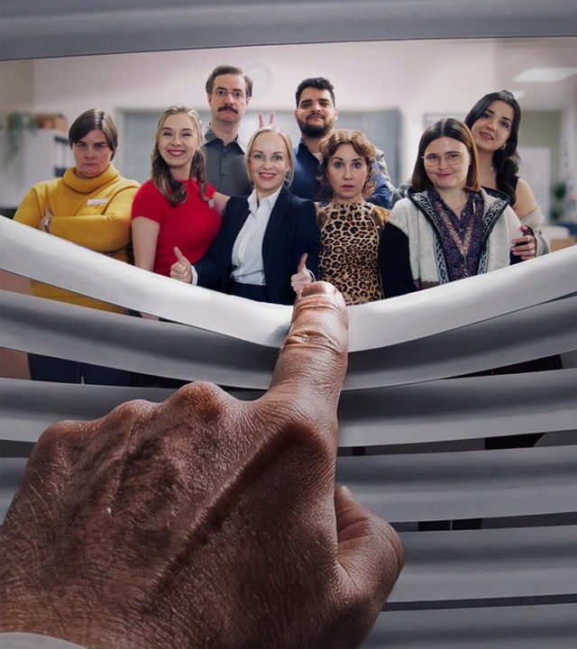 "Nine to Five – Das Amt": Eine Hand biegt eine Jalousie herunter und blickt auf eine Gruppe von sieben lächelnden Büroangestellten. Das diverse Team posiert gemeinsam in einem hellen Großraumbüro.