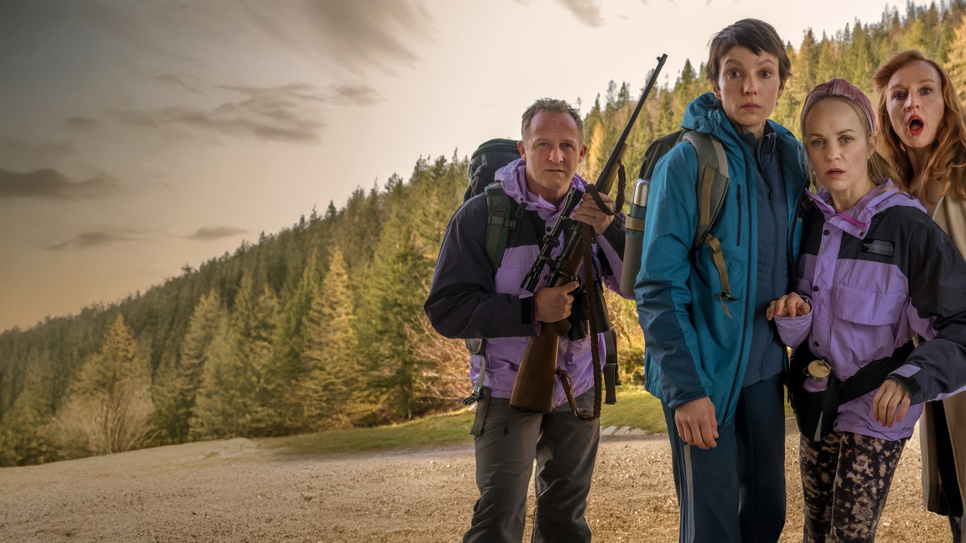 Eine Gruppe von sieben sichtlich schockierten Männern und Frauen blickt in einer Holzhütte gedrängt nach vorne. Eine Frau in der Mitte zielt dabei konzentriert mit einem Gewehr direkt in Richtung der Betrachtenden.