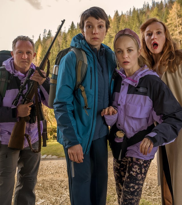 Eine Gruppe von sieben sichtlich schockierten Männern und Frauen blickt in einer Holzhütte gedrängt nach vorne. Eine Frau in der Mitte zielt dabei konzentriert mit einem Gewehr direkt in Richtung der Betrachtenden.