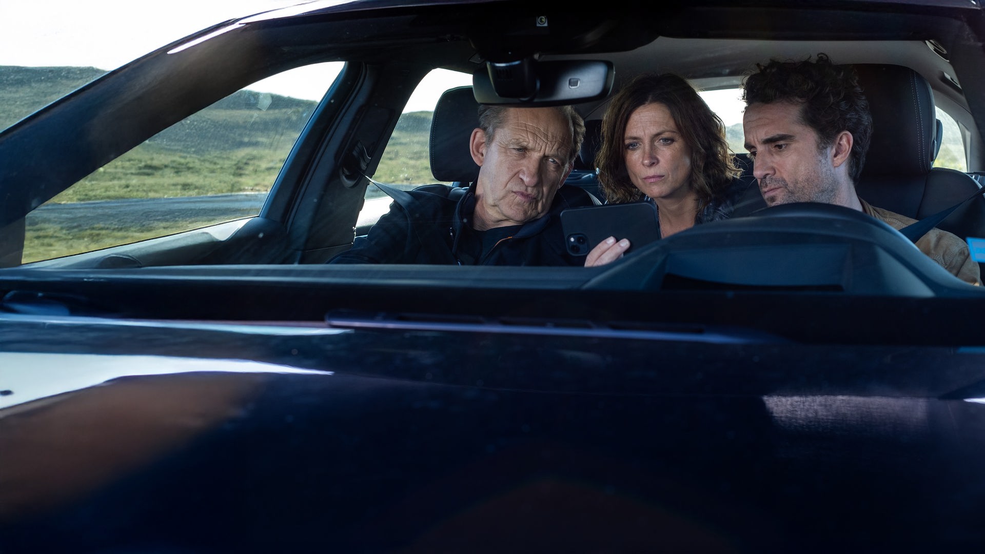 "Nord Nord Mord - Sievers und der Gesang der Wale": Carl Sievers (Peter Heinrich Brix), Ina Behrendsen (Julia Brendler) und Hinnerk Feldmann (Oliver Wnuk) sitzen im Auto – Ina auf der Rückbank – und schauen sich gemeinsam etwas auf Feldmanns Handy an, das er in die Mitte hält. Im Hintergrund liegen die Dünen neben der Straße.