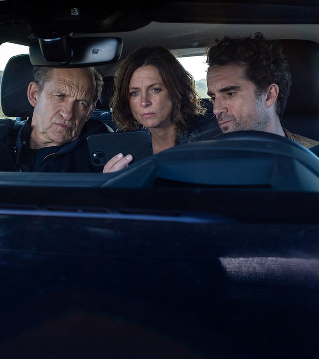 "Nord Nord Mord - Sievers und der Gesang der Wale": Carl Sievers (Peter Heinrich Brix), Ina Behrendsen (Julia Brendler) und Hinnerk Feldmann (Oliver Wnuk) sitzen im Auto – Ina auf der Rückbank – und schauen sich gemeinsam etwas auf Feldmanns Handy an, das er in die Mitte hält. Im Hintergrund liegen die Dünen neben der Straße.