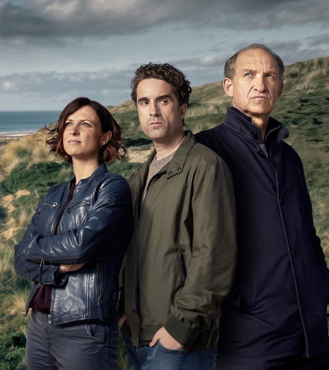 Ina Behrendsen (Julia Bender), Hinnerk Feldmann (Oliver Wnuk) und Karl Sievers (Peter Heinrich Brix),  die Hauotdarsteller der Serie Nord Nord Mord, stehen nebeneinander vor der Kulisse eines typisch norddeutschen Strandes. Direkt im Hintergrund sieht man einen Holzweg durch die grasbewachsenen Dünen am Strand.
