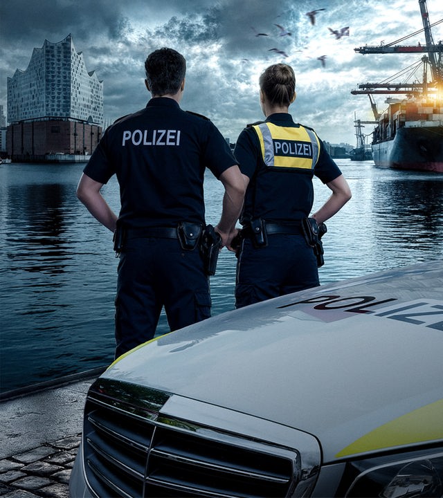 Ein Polizist und eine Polizistin schauen auf das Hafenbecken vor Hamburg. Im Hintergrund ist die Elbphilharmonie zu erkennen. Der Himmel ist grau und wolkenverhangen. Es herrscht eine mysthische Lichtstimmung.