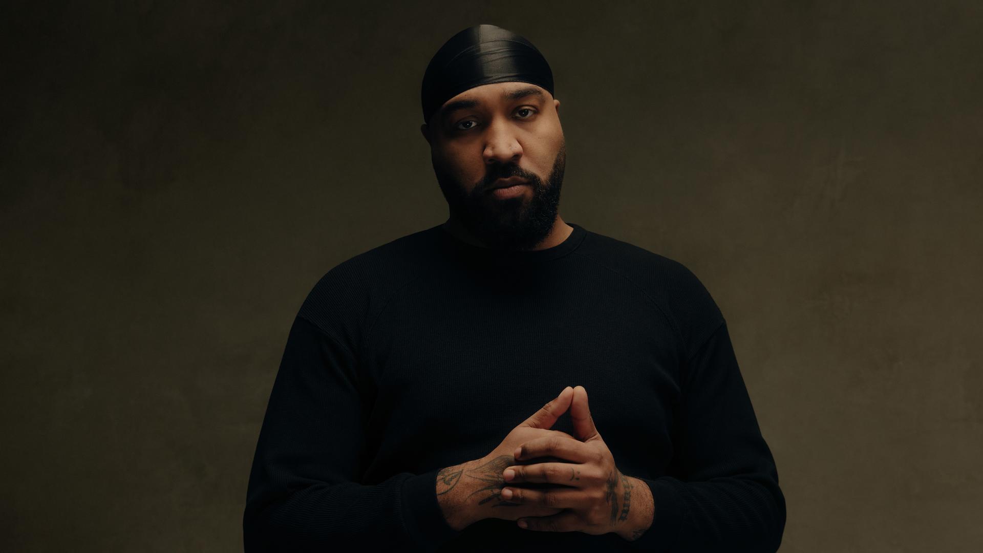 Rapper OG Keemo posiert: Mit gefalteten Händen, schwarzer Kopfbedeckung und schwarzem Oberteil schaut er in Richtung Kamera. er steht vor einem braunen Hintergrund.