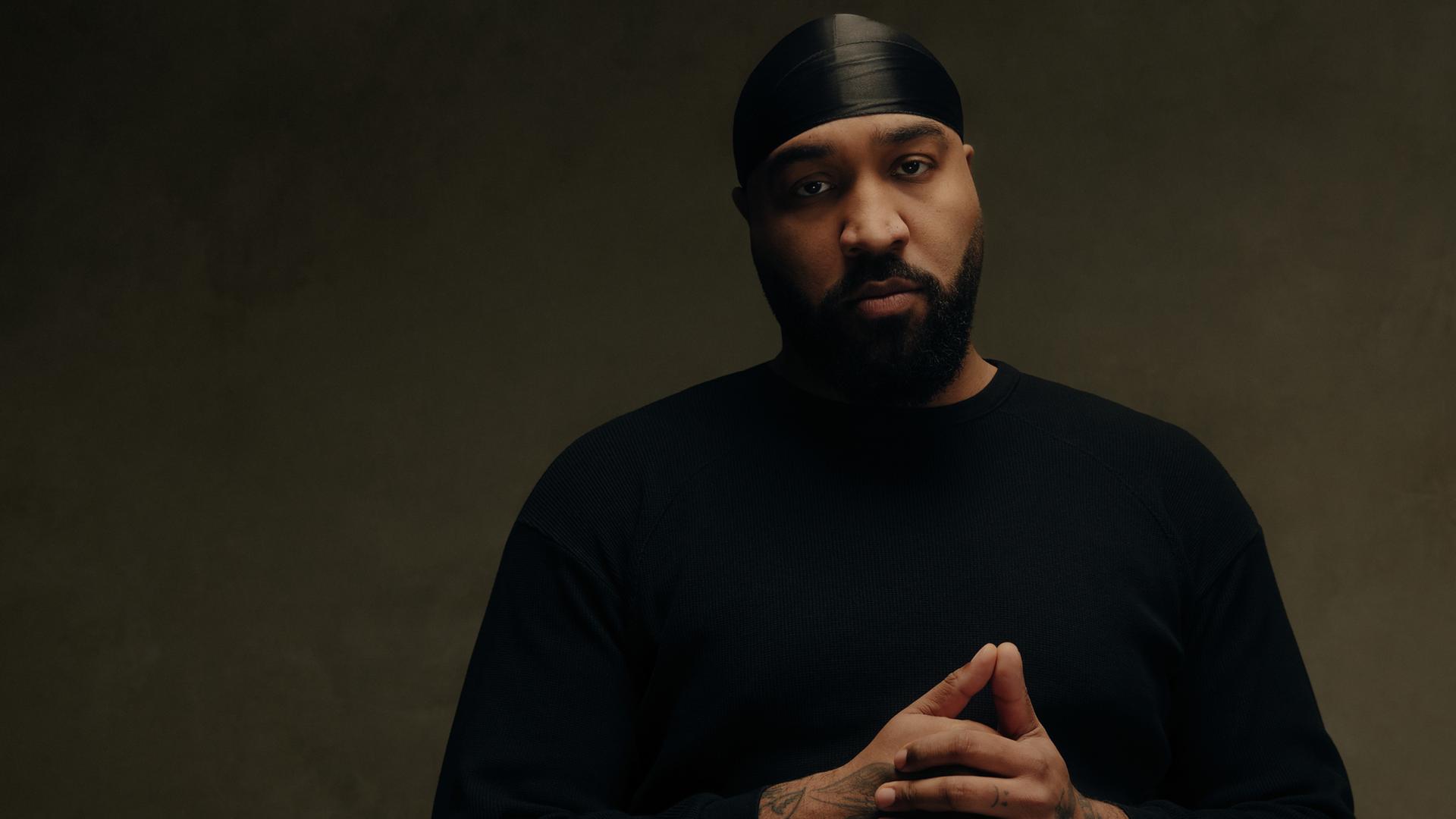 Rapper OG Keemo posiert: Mit gefalteten Händen, schwarzer Kopfbedeckung und schwarzem Oberteil schaut er in Richtung Kamera. Er steht vor einem braunen Hintergrund.