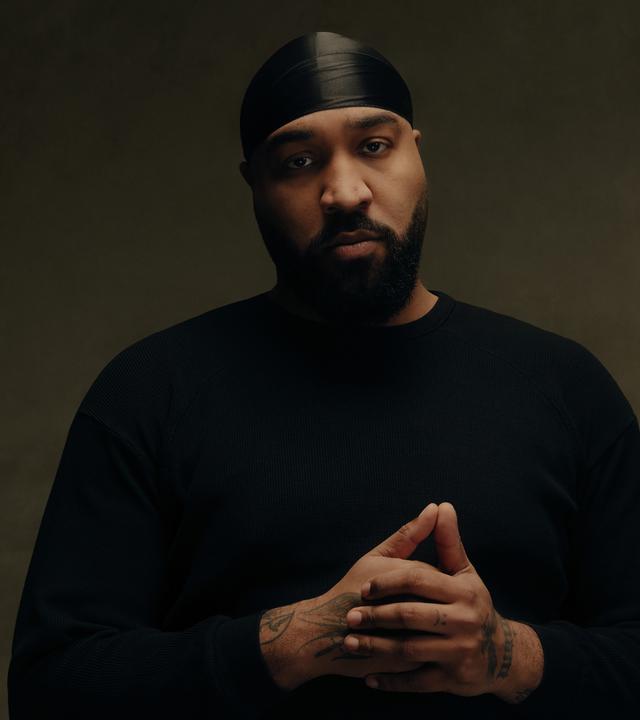 Rapper OG Keemo posiert: Mit gefalteten Händen, schwarzer Kopfbedeckung und schwarzem Oberteil schaut er in Richtung Kamera. Er steht vor einem braunen Hintergrund.