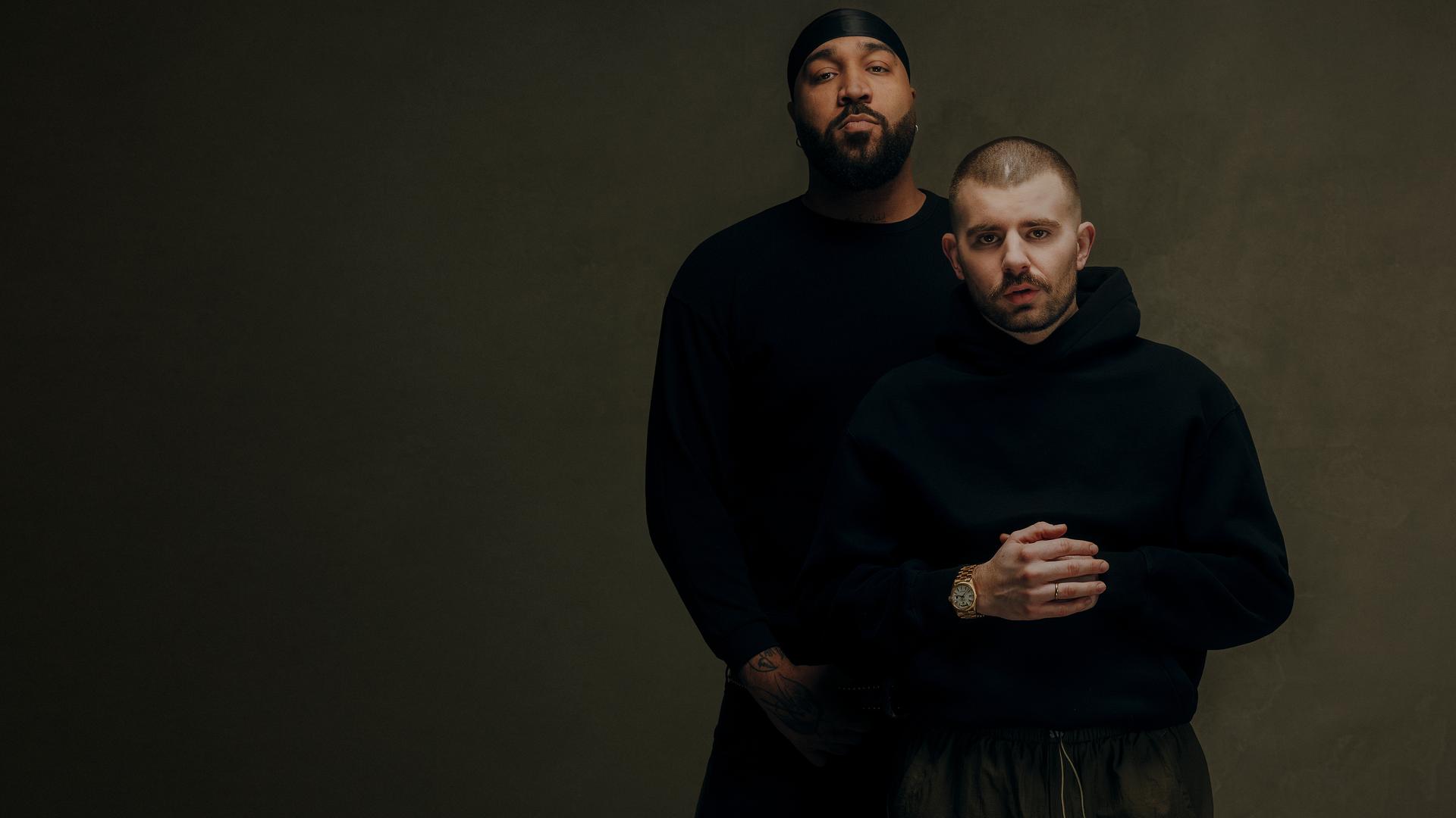 Rapper OG Keemo (links) mit seinem Freund und Produzenten Funkvater Frank. Beide stehen mit schwarzer Kleidung vor einem braunen Hintergrund und schauen in die Kamera.