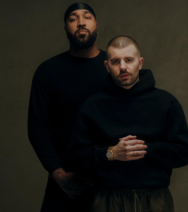 Rapper OG Keemo (links) mit seinem Freund und Produzenten Funkvater Frank. Beide stehen mit schwarzer Kleidung vor einem braunen Hintergrund und schauen in die Kamera.