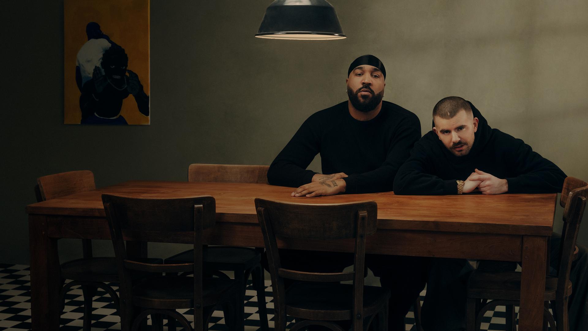 OG Keemo (links) und Funkvater Frank (rechts) sitzen an einem Tisch mit sieben Stühlen und schauen Richtung Kamera geradeaus.