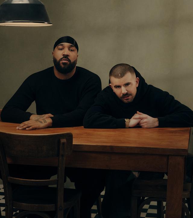 OG Keemo (links) und Funkvater Frank (rechts) sitzen an einem Tisch mit sieben Stühlen und schauen Richtung Kamera geradeaus.