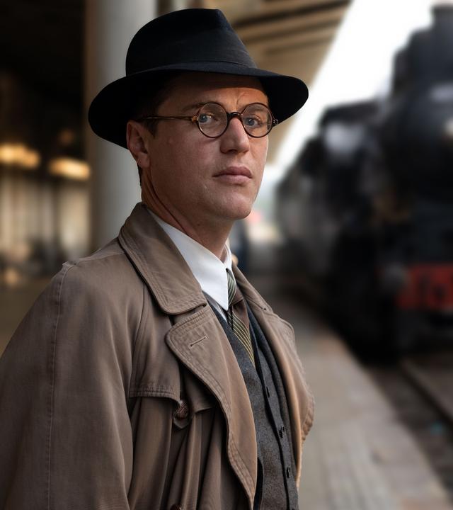 Nicholas (Johnny Flynn) steht im Bahnhof, eine Dampflok fährt ein. Er schaut zur Seite.