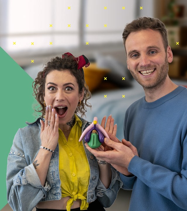 Dr. Leon Windscheid steht im Vordergrund und hält ein farbiges anatomisches Modell einer Klitoris in der Hand. Neben ihm steht Leila Lowfire und blickt überrascht nach vorne. Im Hintergrund befindet sich ein Schlafzimmer mit einem Bett und farbigen Kissen.