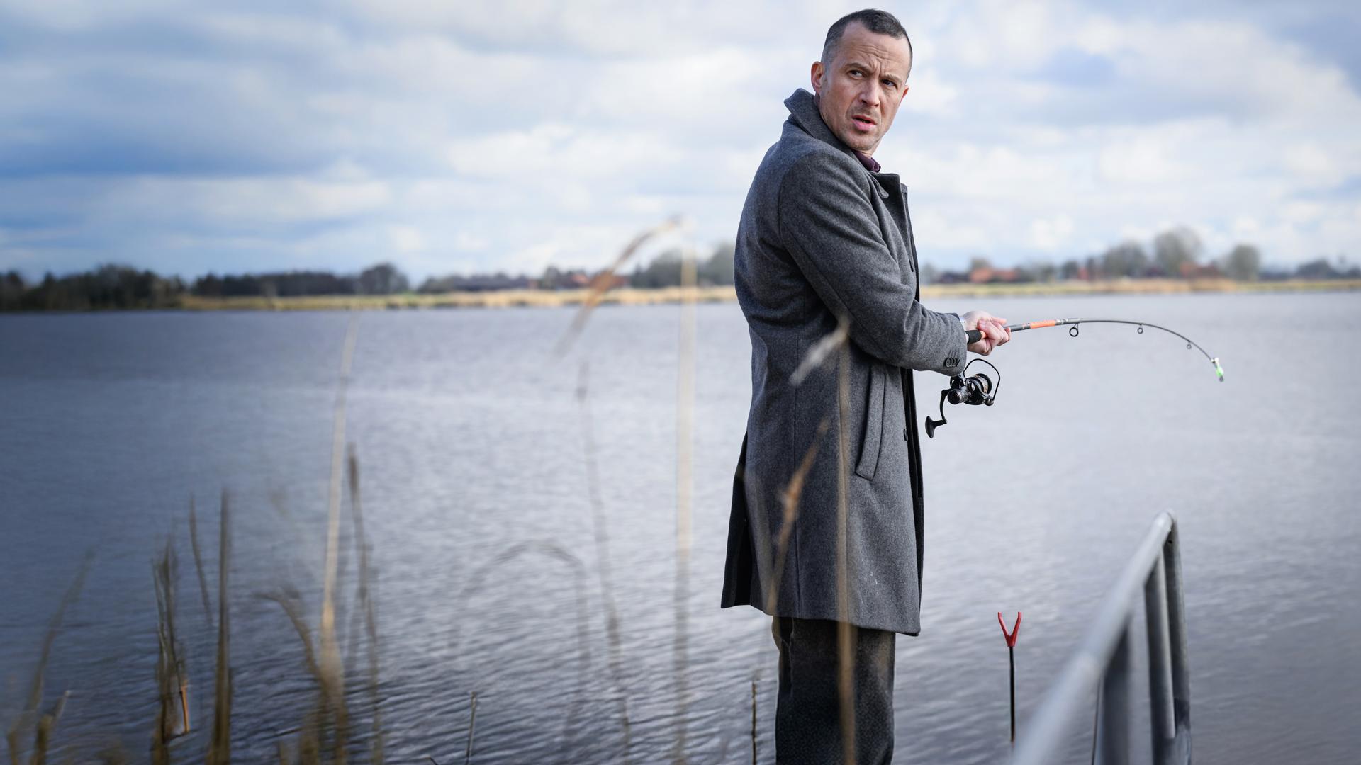 Kommissar Rupert steht am Ufer mit einer Angelrute. er schaut skeptisch über seine Schulter.