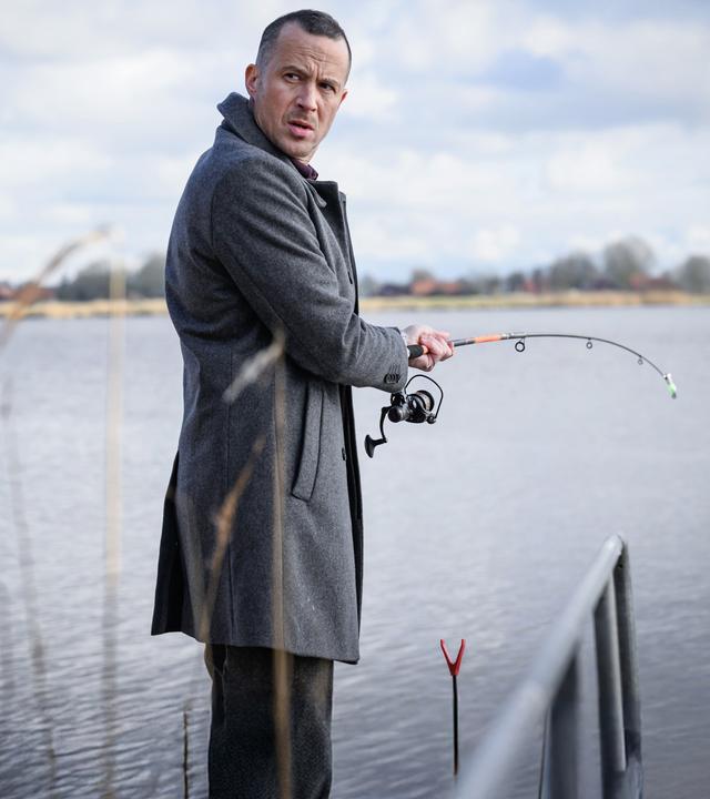 Kommissar Rupert steht am Ufer mit einer Angelrute. Er schaut skeptisch über seine Schulter.
