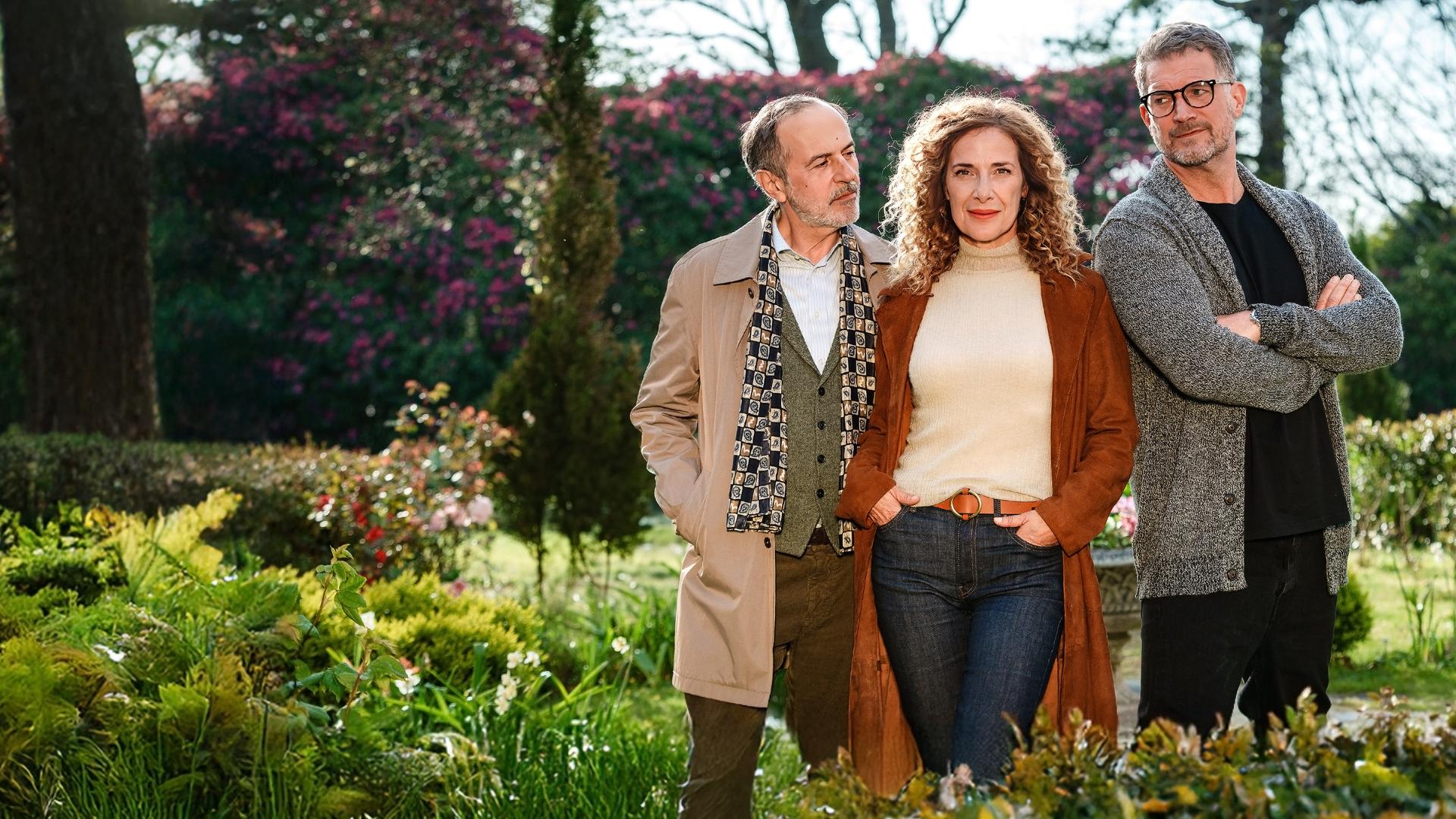Kate Evans (Clelia Sarto) steht in der Mitte zwischen Andrew Evans (Simon Böer) und Yamen Nicholls (Merab Ninidze). Kate hat die Hände in den Hosentaschen und blickt lächelnd in die Kamera.