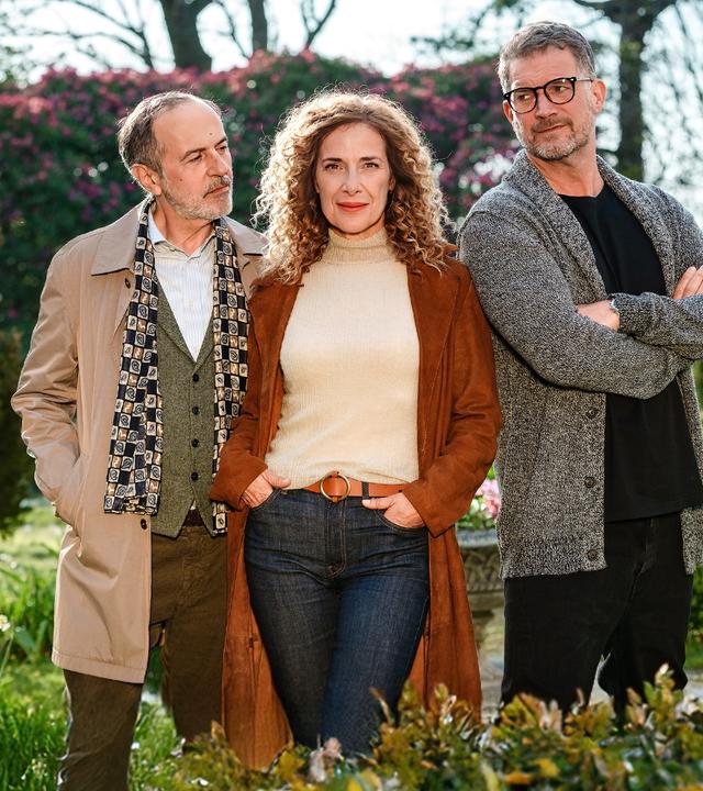 Kate Evans (Clelia Sarto) steht in der Mitte zwischen Andrew Evans (Simon Böer) und Yamen Nicholls (Merab Ninidze). Kate hat die Hände in den Hosentaschen und blickt lächelnd in die Kamera.