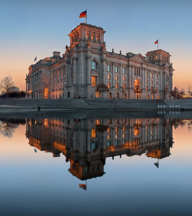 Der Bundestag im Sonnenuntergangslicht. Rechts vom Reichstag ist noch ein Teil des Paul-Löbe-Hauses zu erkennen. Links andere Gebäude im politischen Berlin. Alle Gebäude spiegeln sich am unteren Bildrand in der Spree.