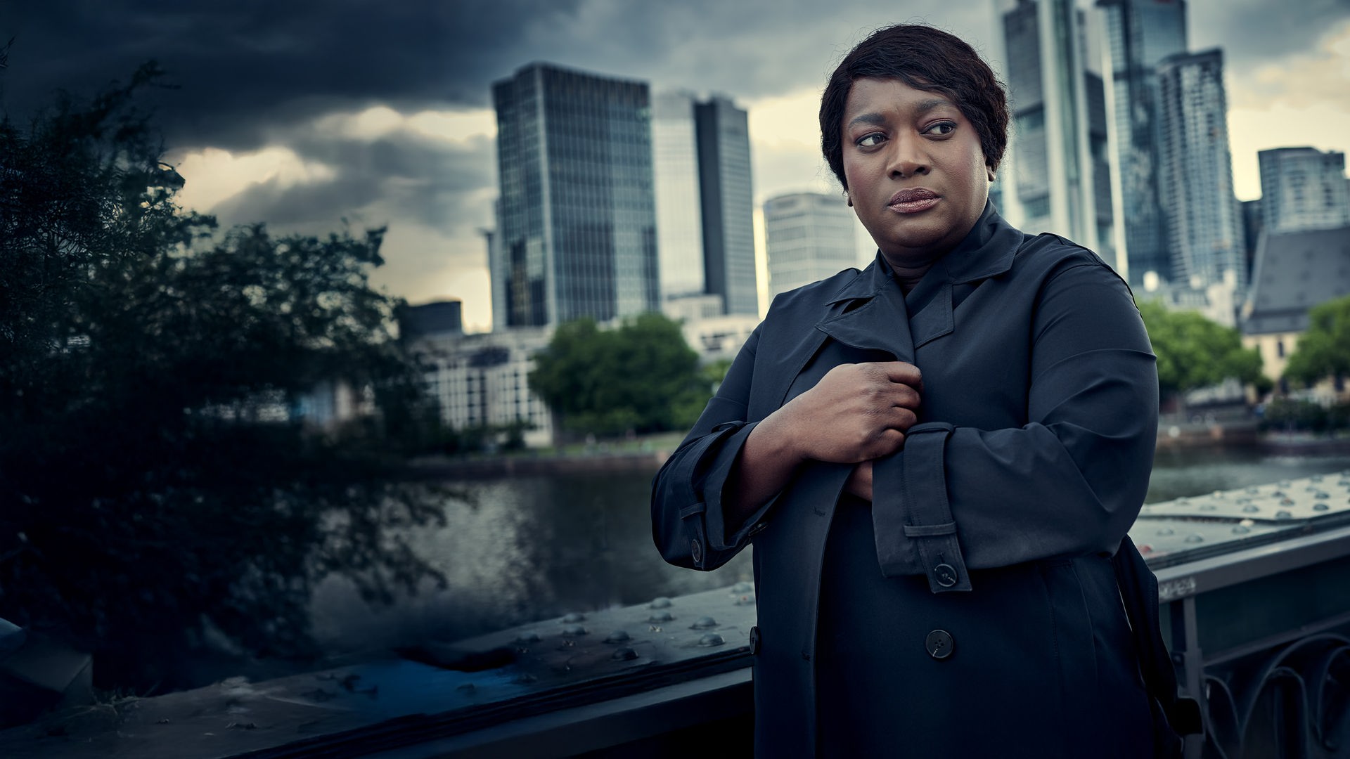 „Die Polizistin und die Todesstille“: Die BKA-Fallanalystin Gloria Acheampong (Thelma Buabeng) im dunklen Mantel vor der Skyline von Frankfurt am Main. Sie blickt mit ernstem, nachdenklichem Gesichtsausdruck leicht zur Seite, während im Hintergrund markante Hochhäuser unter einem bewölkten Himmel stehen.