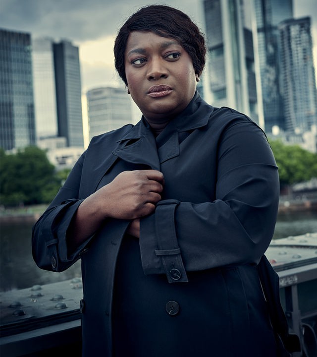 „Die Polizistin und die Todesstille“: Die BKA-Fallanalystin Gloria Acheampong (Thelma Buabeng) im dunklen Mantel vor der Skyline von Frankfurt am Main. Sie blickt mit ernstem, nachdenklichem Gesichtsausdruck leicht zur Seite, während im Hintergrund markante Hochhäuser unter einem bewölkten Himmel stehen.