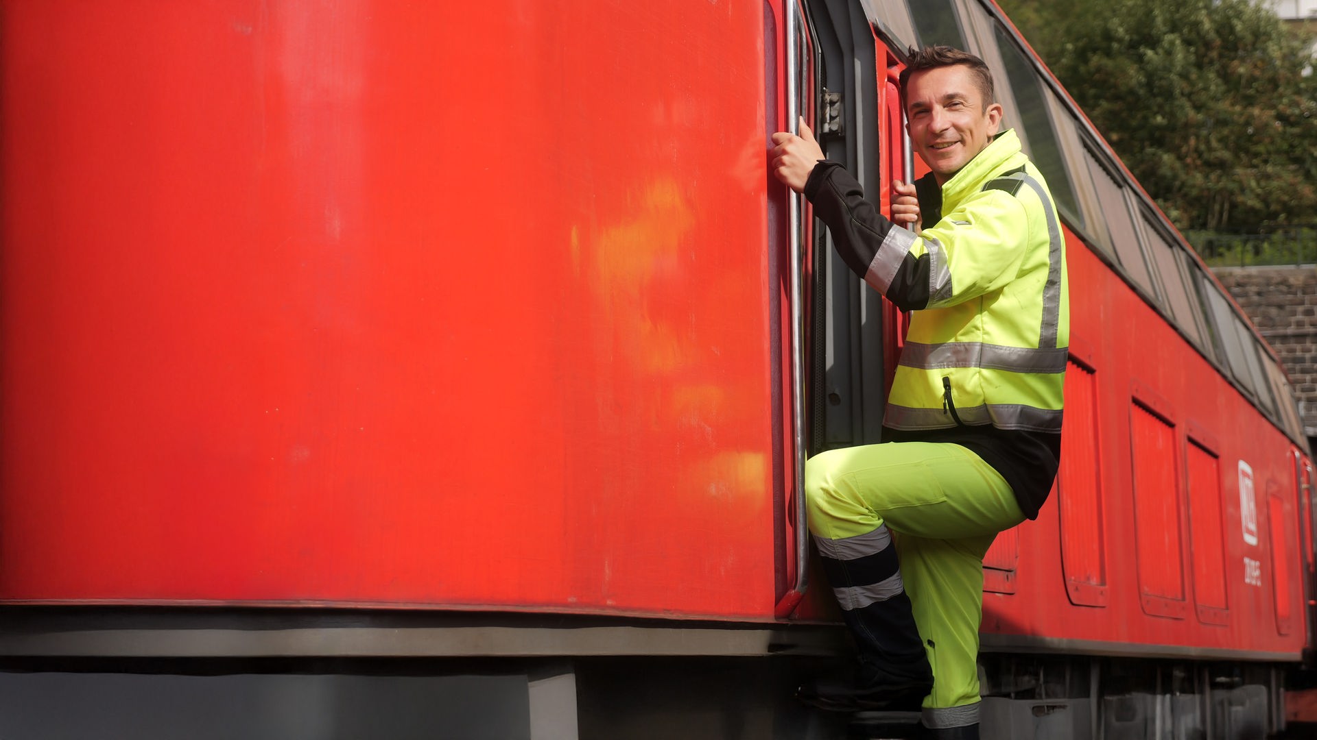 Eric Mayer erklimmt in einer gelben Arbeitsmontur die Leiter zum Führerstand einer roten Lokomotive. Dabei lächelt er in die Kamera.