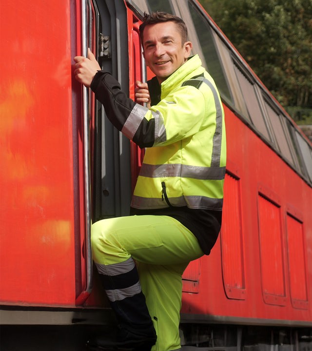 Eric Mayer erklimmt in einer gelben Arbeitsmontur die Leiter zum Führerstand einer roten Lokomotive. Dabei lächelt er in die Kamera.