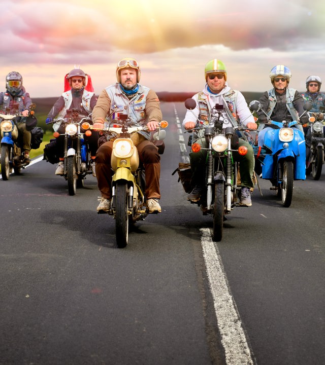 Vier Männer fahren auf Simson-Mopeds auf einer Landstraße in V-Formation auf die Kamera zu. Die Landschaft, die im Hintergrund zu sehen ist, ist KI-generiert. 