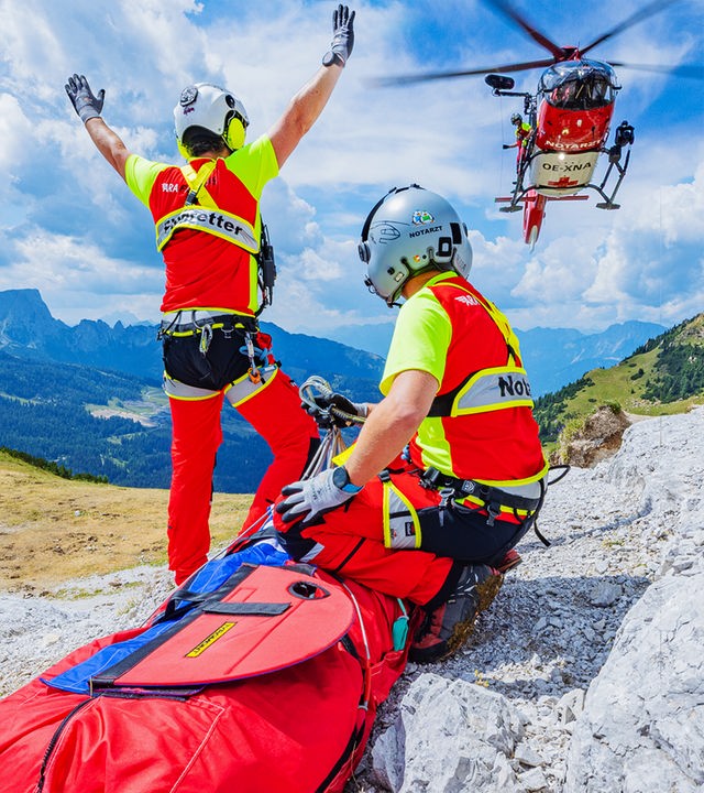 Alpenpanorama: Im Vordergrund warten zwei Flugretter mit Rettungssack auf den im Hintergrund einschwebenden Helikopter.