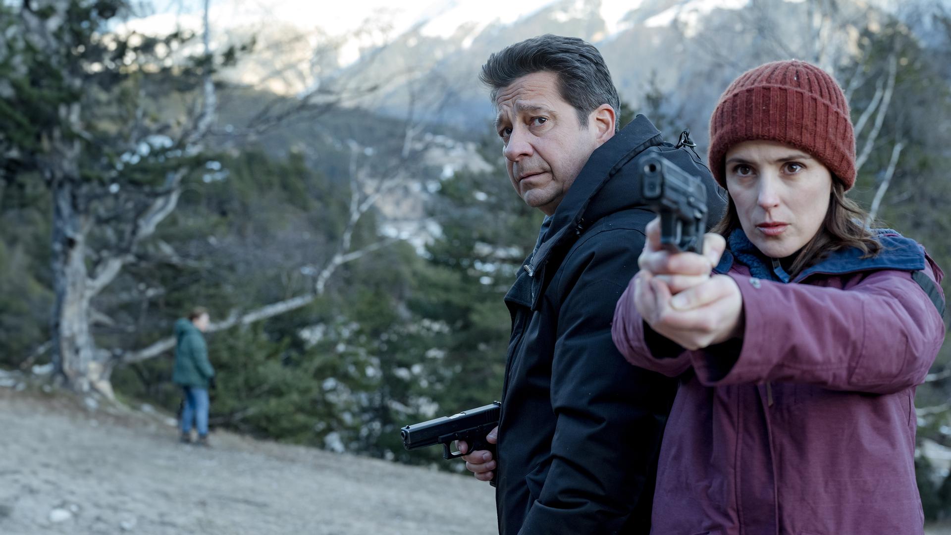 Andréas (Laurent Gerra) und Constance (Clémentine Poidatz) stehen tagsüber in einer winterlichen Berglandschaft. Constance richtet eine Waffe seitlich nach vorne. Neben ihr blickt Andréas ebenfalls angespannt nach vorne.